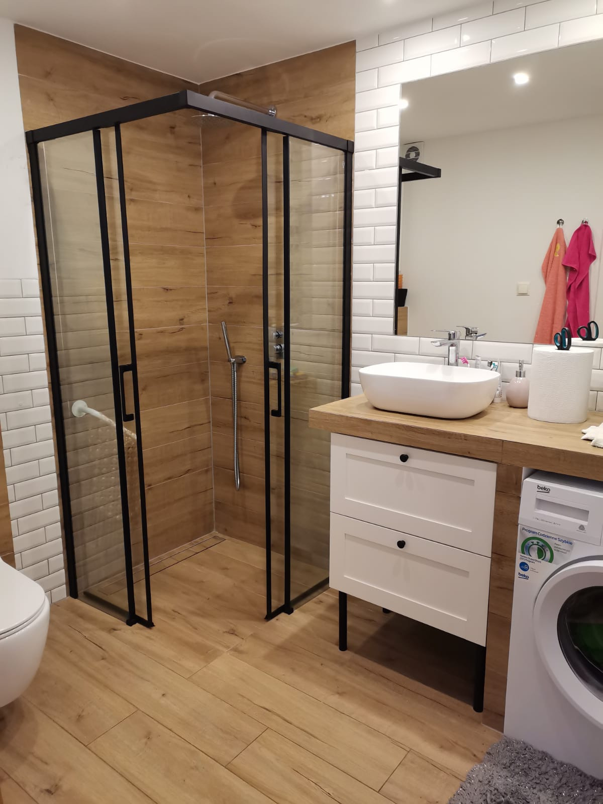 Nowoczesna łazienka z kabiną prysznicową typu walk-in z czarnymi ramami, białą umywalką nablatową i drewnopodobnymi płytkami. Widoczna pralka i białe płytki na ścianach.