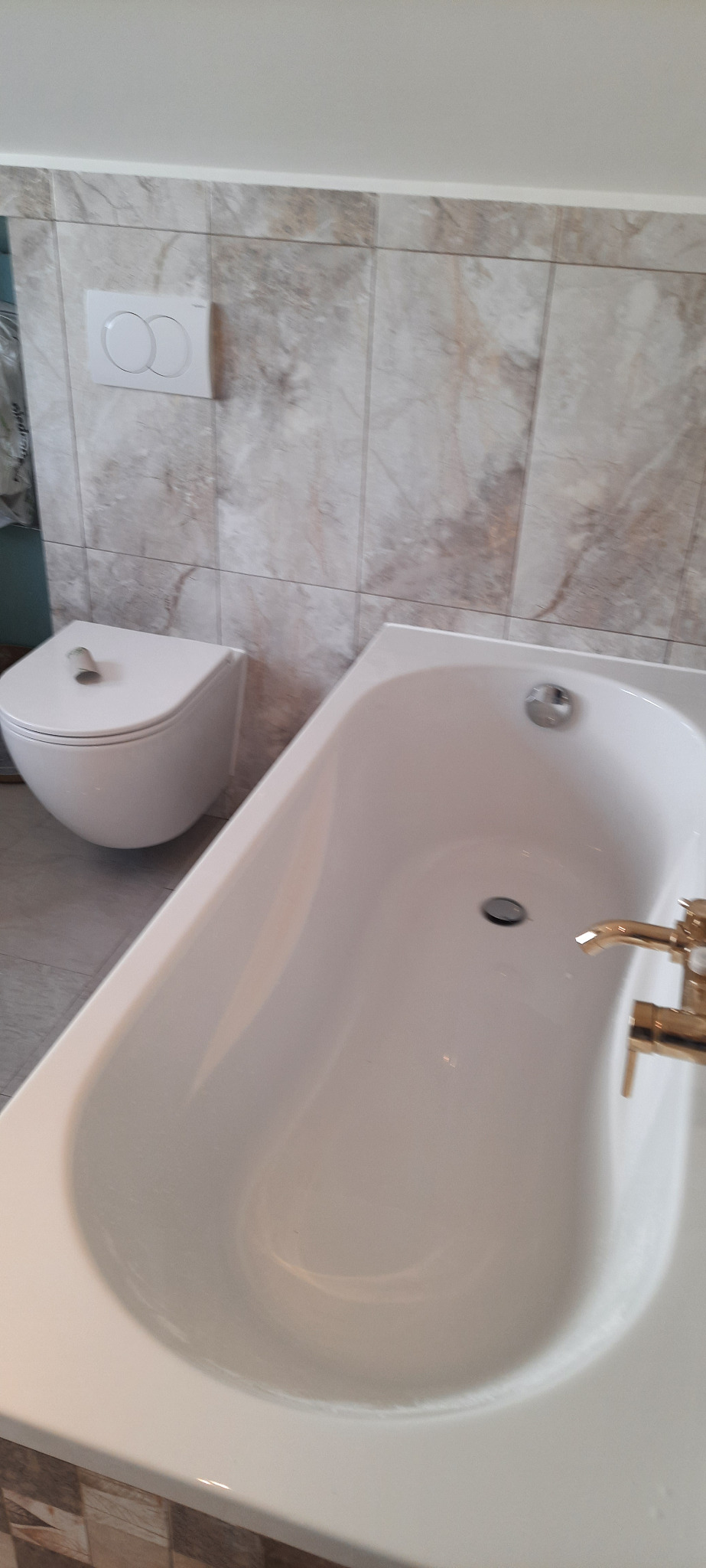 Biała wanna z baterią w kolorze złota i podwieszana toaleta na tle płytek imitujących marmur. Wykończona łazienka po remoncie.