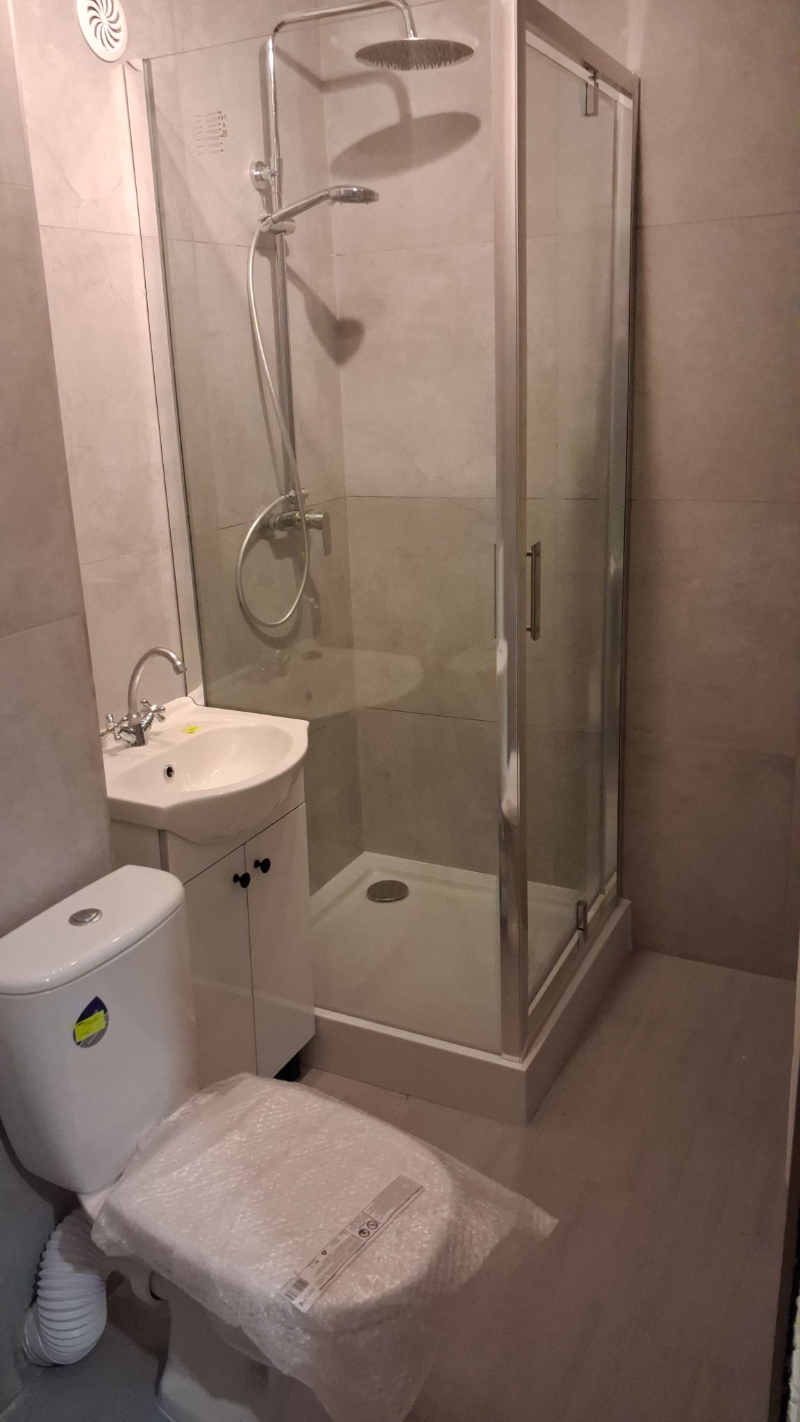 Wykończona łazienka w beżowych płytkach z kabiną prysznicową, umywalką z szafką i toaletą z folią ochronną na desce. Nowoczesny design, widok z bliska.