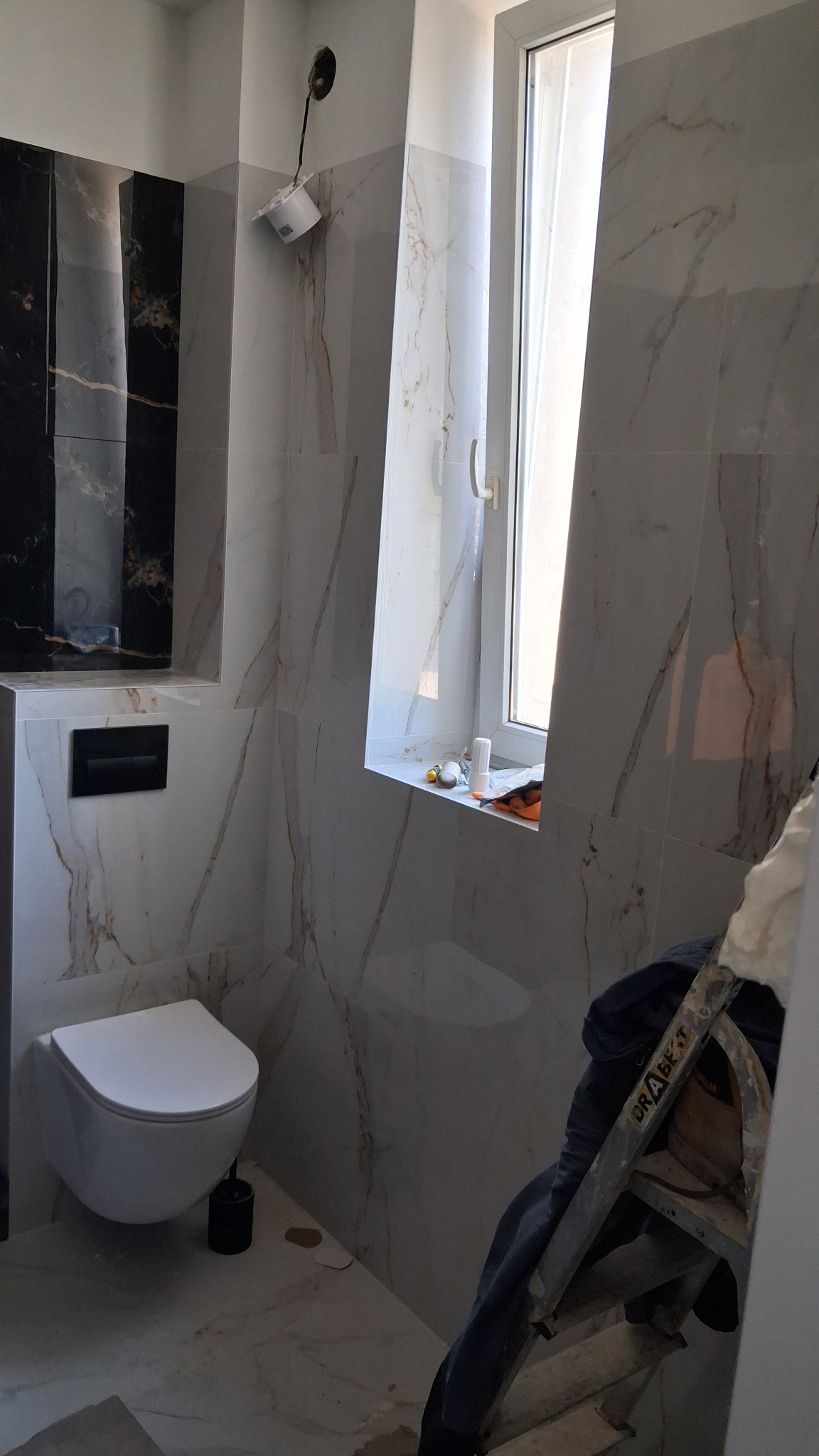 Wykończona łazienka z marmurowymi płytkami, wiszącą toaletą i oknem. Widoczna drabina sugeruje trwające prace wykończeniowe. Czarno-biała kolorystyka płytek.