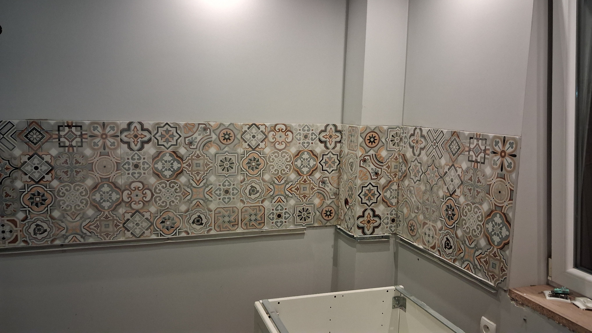 Wnętrze w trakcie wykańczania: fragment ściany z dekoracyjnymi płytkami ceramicznymi w stylu patchwork, widoczne profile montażowe i fragment szafki kuchennej.