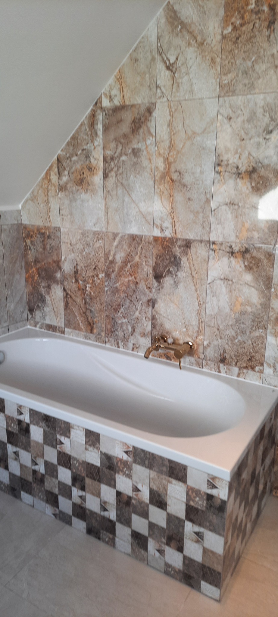 Wykończona łazienka na poddaszu: biała wanna obudowana mozaiką, ściany wyłożone płytkami imitującymi marmur, złota bateria. Ukośny sufit.