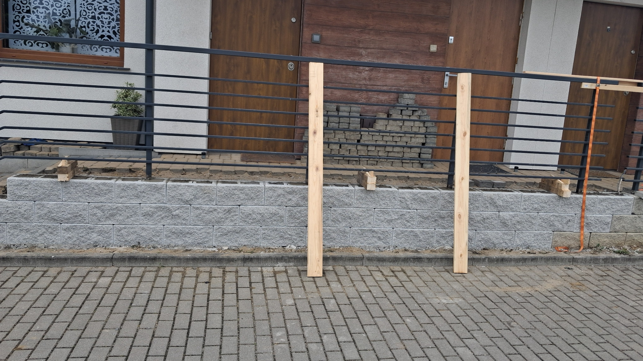 Wznoszenie murku oporowego z szarej kostki betonowej. Widoczne drewniane podpory, czarna balustrada i ułożona kostka brukowa. Etap budowy przed domem.