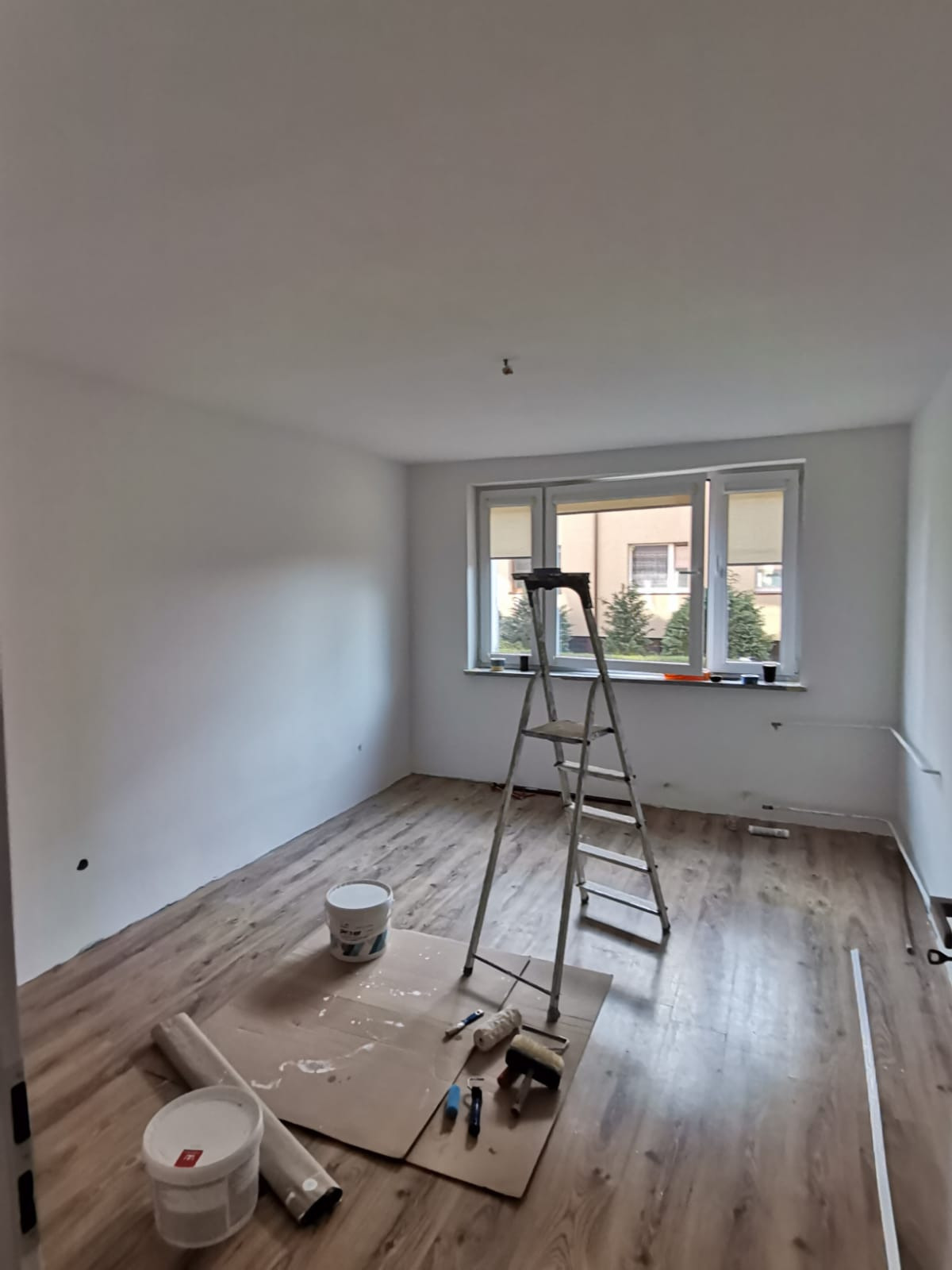 Pokój po malowaniu ścian na biało, z drabiną, farbami i narzędziami malarskimi na podłodze z paneli. Widok na okno z zewnątrz.