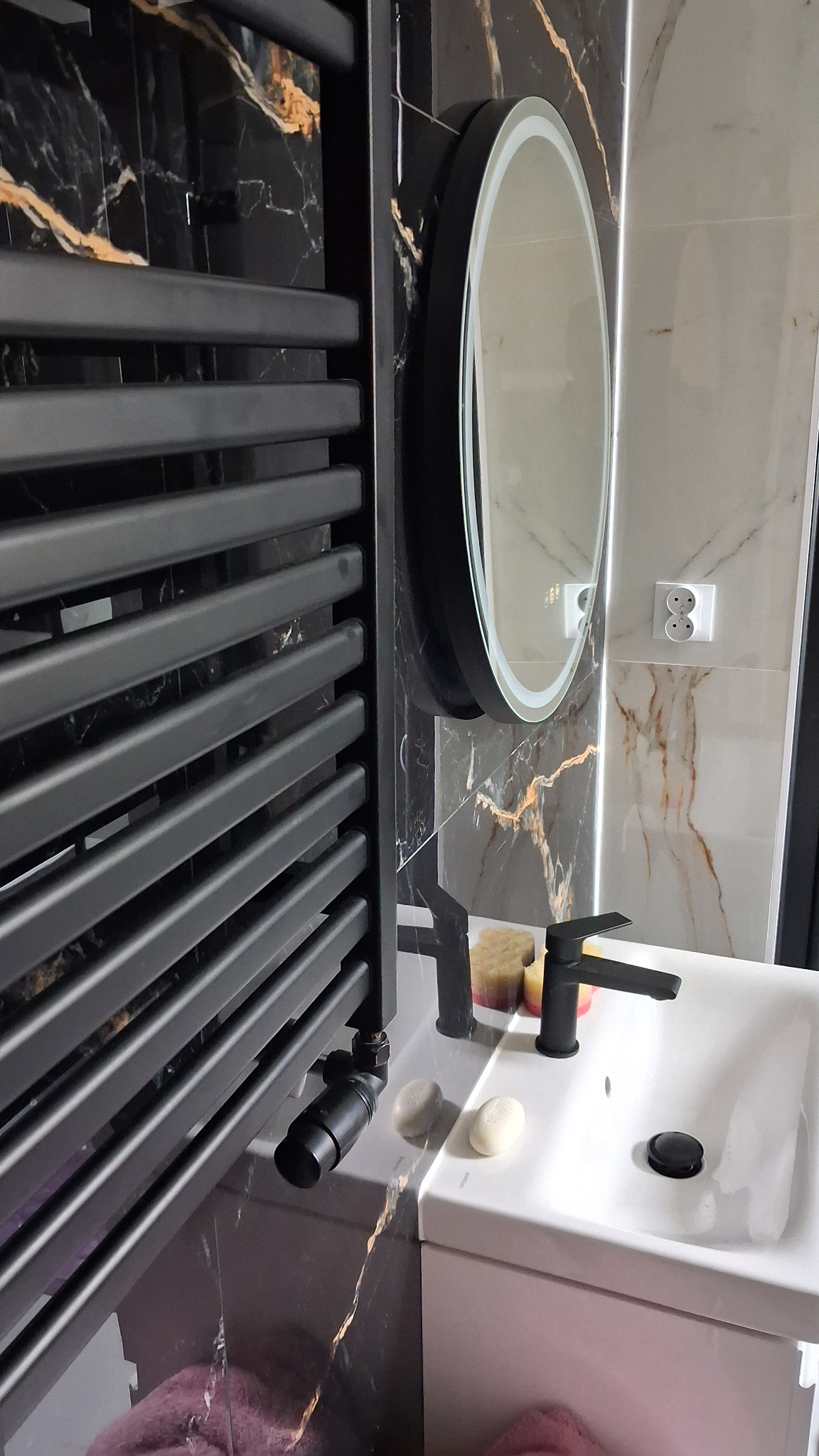 Nowoczesna łazienka z czarnym grzejnikiem drabinkowym, owalnym lustrem z podświetleniem LED i umywalką nablatową na tle marmurowych płytek.