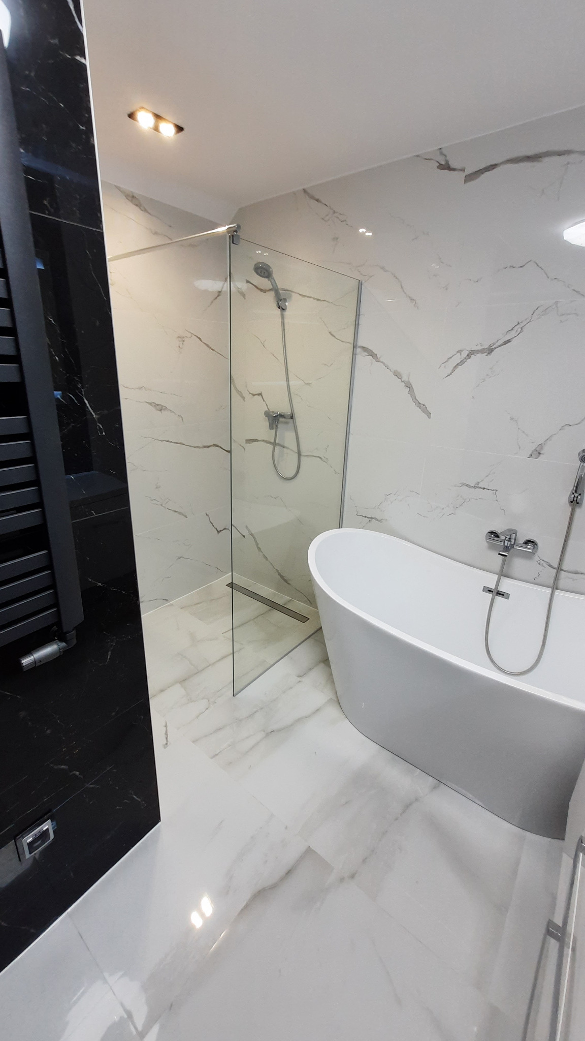 Nowoczesna łazienka z kabiną prysznicową typu walk-in i wolnostojącą wanną, wykończona białym marmurem z szarymi żyłami. Czarny grzejnik drabinkowy.