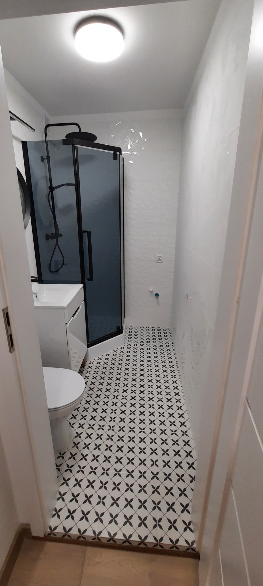 Nowoczesna łazienka z kabiną prysznicową typu walk-in z czarnymi detalami, białą umywalką i toaletą. Na podłodze wzorzyste, czarno-białe płytki.