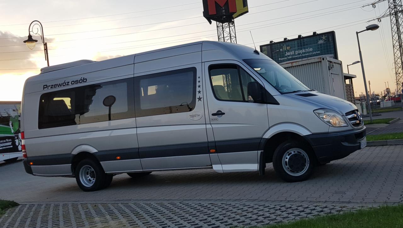 Srebrny bus Mercedes-Benz Sprinter z napisem na oknie 'Przewóz osób' i pięcioma gwiazdkami, zaparkowany na kostce brukowej, w tle słup oświetleniowy i billboard.