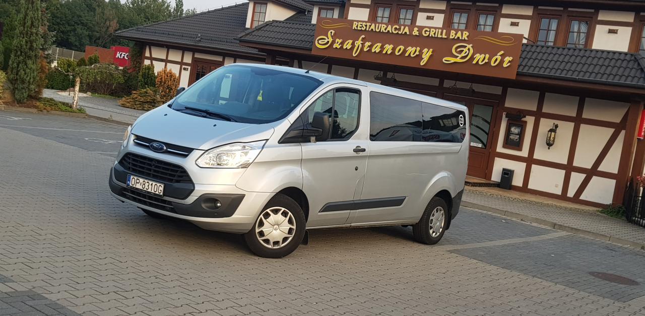 Srebrny Ford Transit Custom z przodu restauracji Szafranowy Dwór, prezentujący usługi transportowe z dbałością o komfort podróży.
