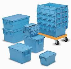 Stos niebieskich, plastikowych pojemników transportowych, niektóre z pokrywami, jeden stos na żółtym wózku z kółkami, w jasnym otoczeniu.