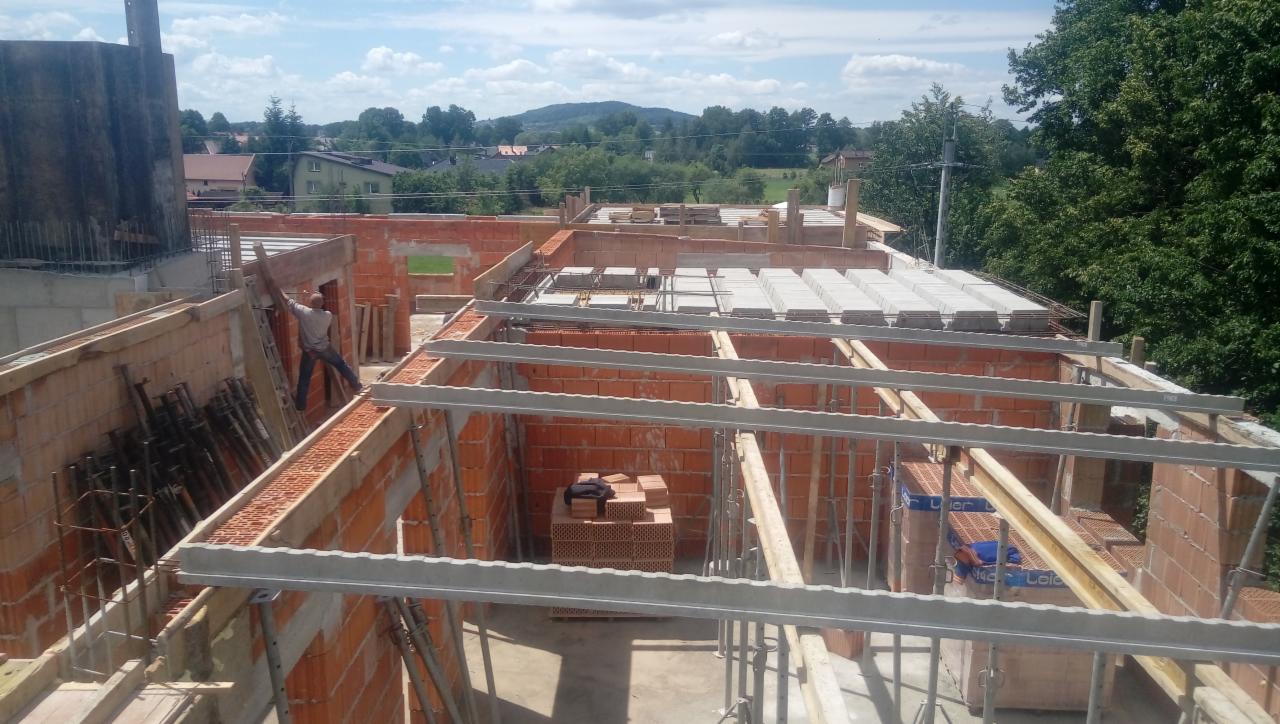 Wznoszenie ścian z czerwonej cegły na budowie domu jednorodzinnego, widoczne stropy betonowe i stalowe elementy konstrukcyjne, pracownik na drabinie w tle.