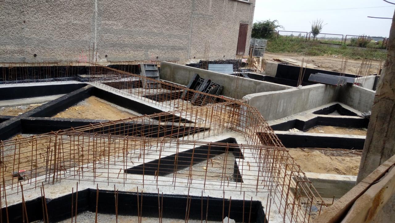 Zalane fundamenty z hydroizolacją i zbrojeniem na placu budowy, widoczne szalunki i elementy konstrukcyjne.