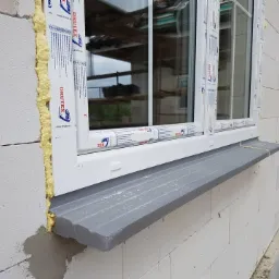 Montaż okna PCV z szarym parapetem z konglomeratu w surowym murze, widoczna pianka montażowa i folie ochronne na ramie okiennej.