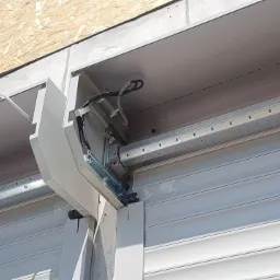 Szczegółowe ujęcie mechanizmu i montażu zewnętrznej rolety okiennej, widoczne prowadnice, zwinięte lamele i obudowa, na tle surowej ściany budynku.