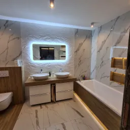 Nowoczesna łazienka z marmurowymi płytkami, podświetlanym lustrem LED, dwiema umywalkami nablatowymi i wanną obudowaną drewnopodobnymi panelami, podświetlona listwą LED przy podłodze.