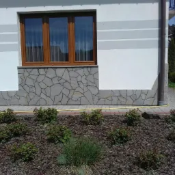 Fragment elewacji domu z oknem, wykończony kamieniem dekoracyjnym, z widocznym fragmentem ogrodu z korą i niskimi krzewami.