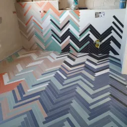 Wnętrze łazienki w trakcie remontu z ułożonymi kolorowymi płytkami w jodełkę na podłodze i częściowo na ścianie, widoczne instalacje wodne i surowe ściany.