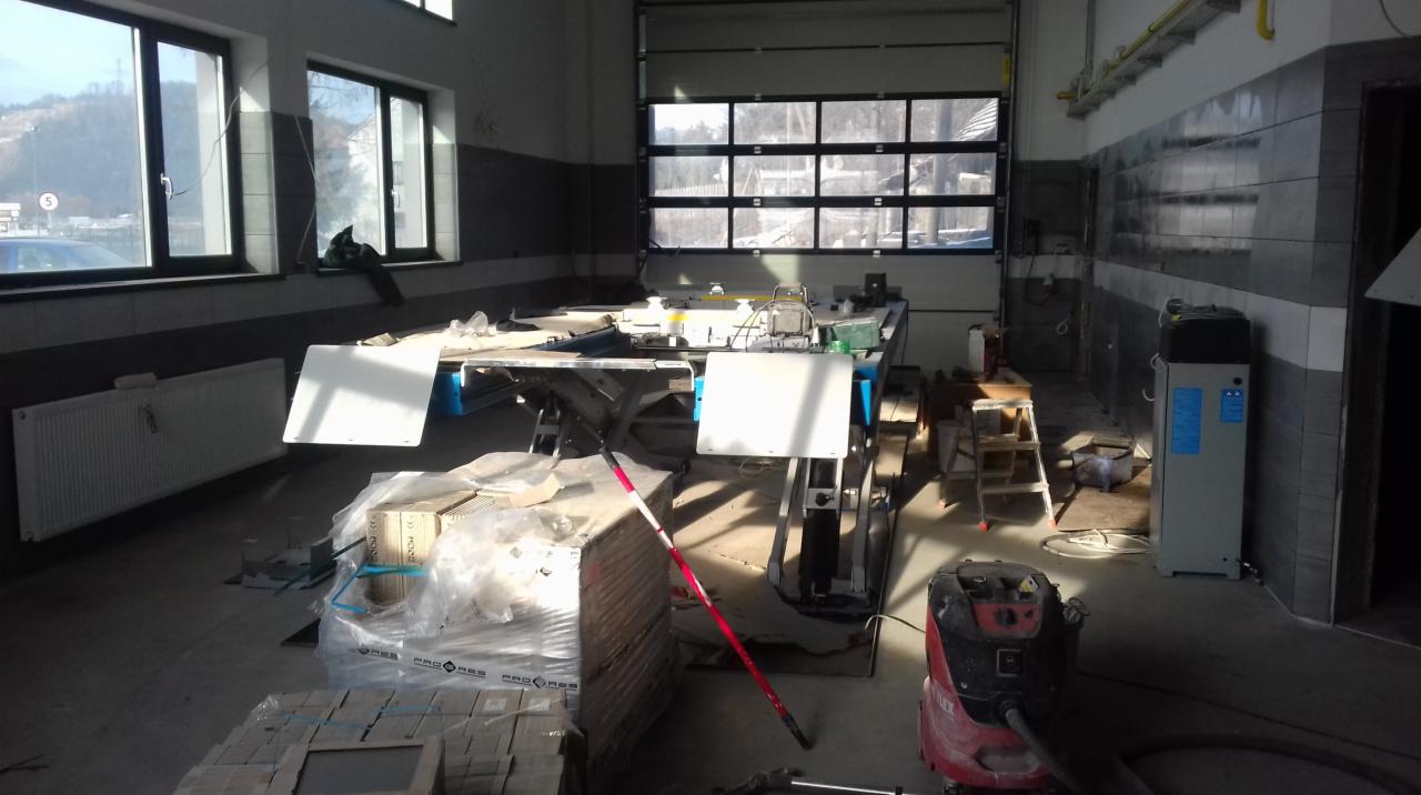 Wnętrze remontowanego garażu z podnośnikiem samochodowym, z widocznymi płytkami na ścianach, materiałami budowlanymi i narzędziami pracy, w trakcie prac wykończeniowych.