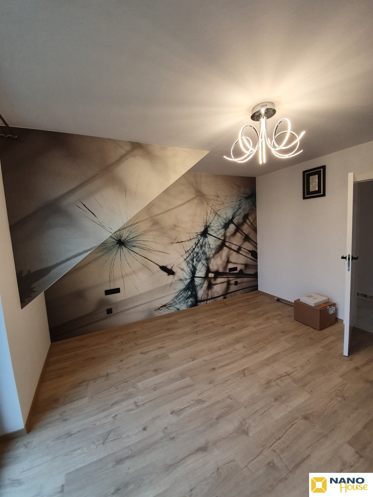 Pokój po remoncie z fototapetą przedstawiającą dmuchawca na skośnej ścianie poddasza, nowoczesna lampa sufitowa, panele podłogowe w odcieniach drewna.