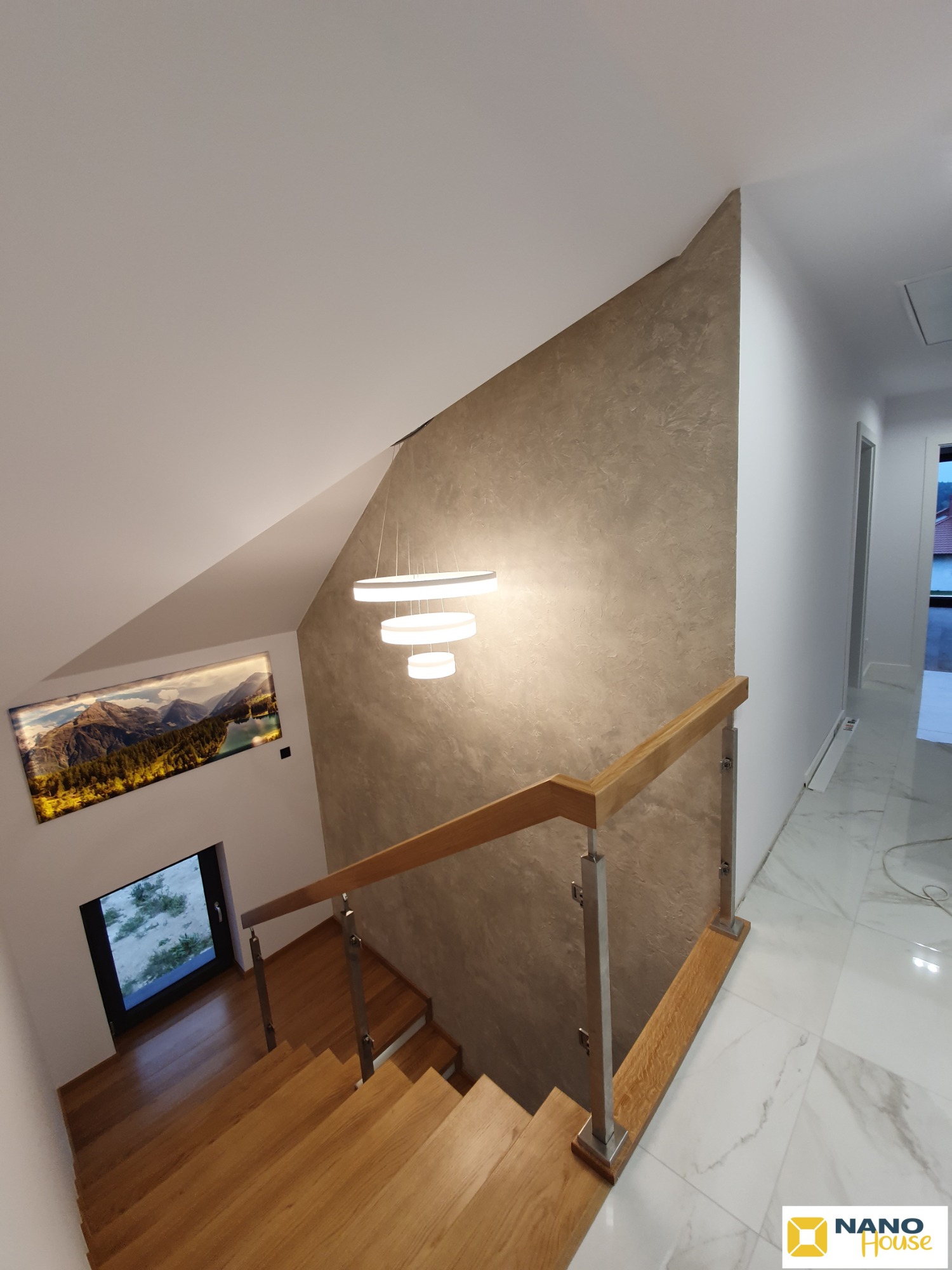 Klatka schodowa w domu z drewnianymi schodami i balustradą ze stali nierdzewnej, ściana wykończona dekoracyjnym tynkiem w odcieniach beżu, okno na podeście, nowoczesna lampa wisząca z trzema okręgami.