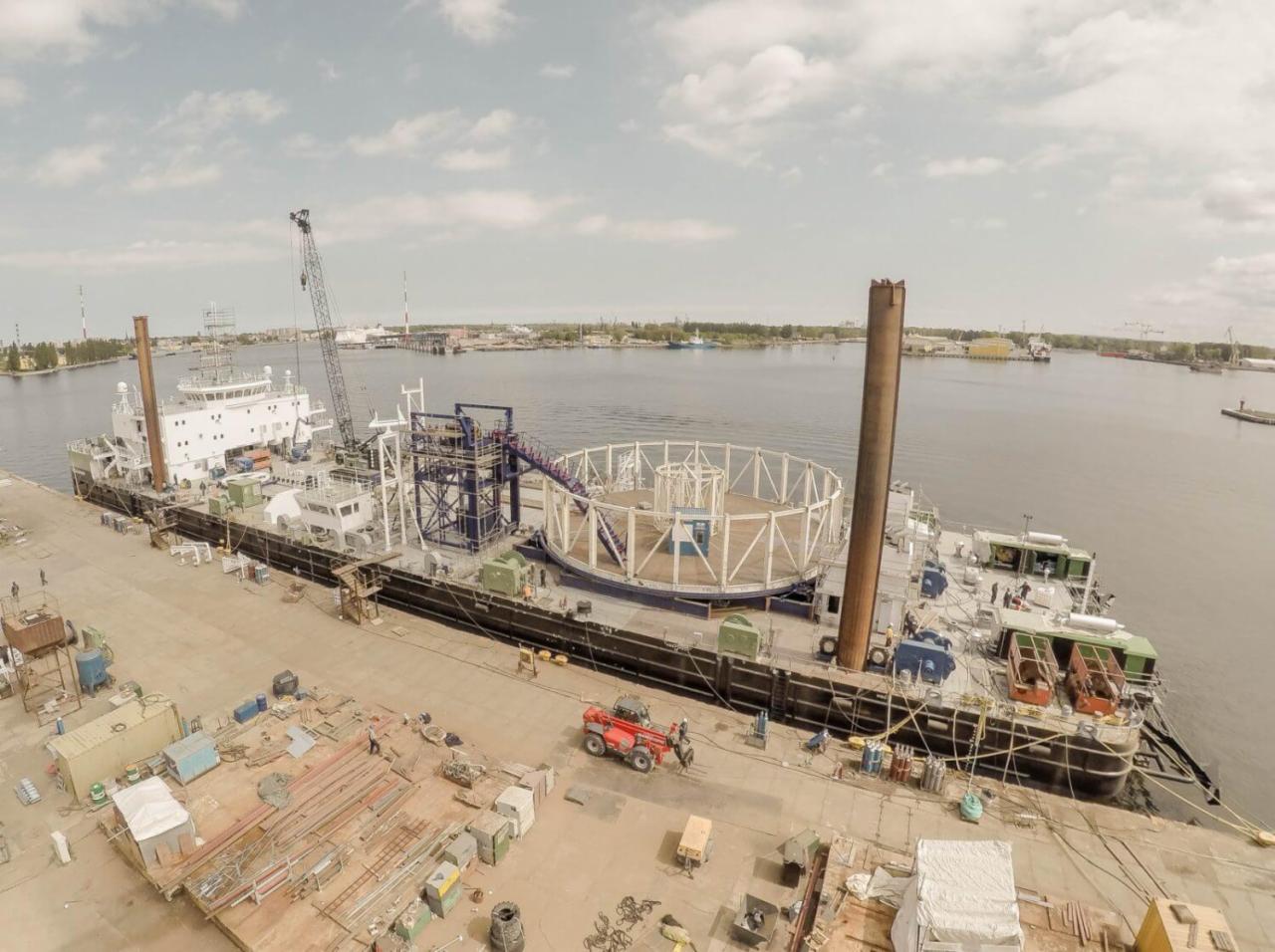 Widok z góry na barkę z konstrukcją stalową w kształcie cylindra i palem wbitym w pokład, na nabrzeżu portowym; w tle widoczne budynki portowe i dźwig.