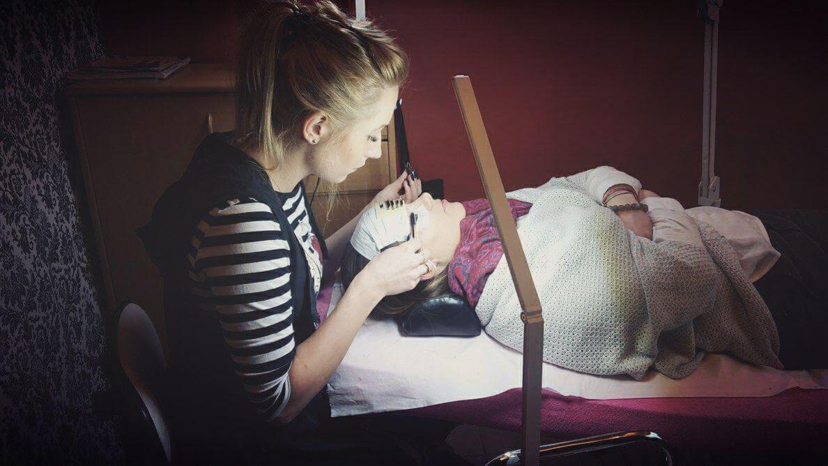 Stylistka rzęs aplikuje sztuczne rzęsy klientce leżącej na kozetce pod lampą, widoczna paletka rzęs na czole klientki.