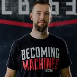 Portret mężczyzny z brodą, ubranego w czarną koszulkę z napisem 'BECOMING MACHINES SINCE 28', na tle szarej ściany z białymi napisami i czerwonym akcentem.