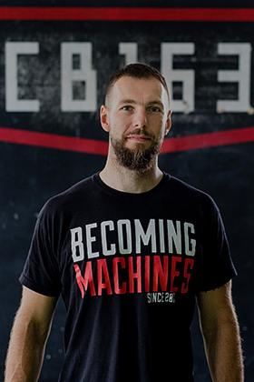 Portret mężczyzny z brodą, ubranego w czarną koszulkę z napisem 'BECOMING MACHINES SINCE 28', na tle szarej ściany z białymi napisami i czerwonym akcentem.