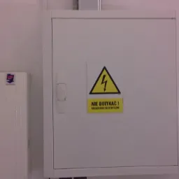 Biała skrzynka elektryczna z żółtą naklejką ostrzegawczą 'NIE DOTYKAĆ! Urządzenie elektryczne' i symbolem błyskawicy w trójkącie.