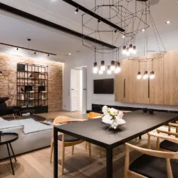 Nowoczesne wnętrze salonu z ceglaną ścianą, szarą sofą, czarnym stołem jadalnym z drewnianymi krzesłami i geometryczną lampą sufitową.