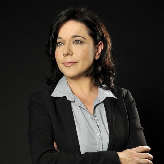 Portret kobiety w czarnej marynarce i szarej koszuli na ciemnym tle. Profesjonalny wizerunek osoby z delikatnym uśmiechem i spojrzeniem w bok, sugerujący zaufanie.