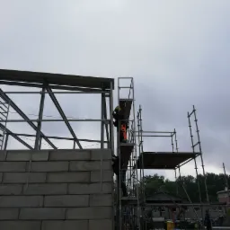 Montaż konstrukcji stalowej hali z użyciem rusztowania, widoczny fragment ściany z betonowych bloczków i pochmurne niebo.