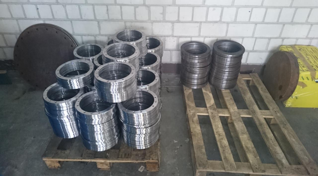 Stosy metalowych pierścieni z otworami na śruby, ułożone na drewnianych paletach, z metalowymi pokrywami w tle, na tle ceglanej ściany.