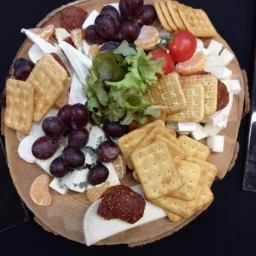 Deska serów z winogronami, krakersami, mandarynkami, pomidorami koktajlowymi i sałatą na drewnianym talerzu na czarnym tle.