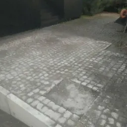 Fragment powierzchni wyłożonej szarą kostką brukową, z widocznymi brakami w ułożeniu i śladami zaprawy, obok niskiego betonowego obrzeża i taczki z pomarańczowym kołem w tle.