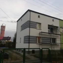 Dwu kondygnacyjny dom z nową białą elewacją z szarymi pasami, częściowo zasłonięty rusztowaniem, w tle widoczna wieża ciśnień.