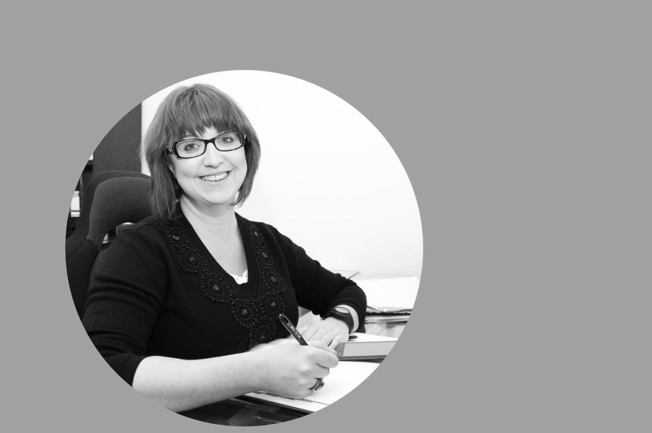 Czarno-białe zdjęcie portretowe uśmiechniętej kobiety w okularach, piszącej długopisem na kartce na biurku, widoczny zegarek na ręku i książka w tle.