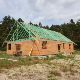 Surowy stan domu murowanego z cegły ceramicznej z widoczną więźbą dachową z zielonych łat, w tle las iglasty.