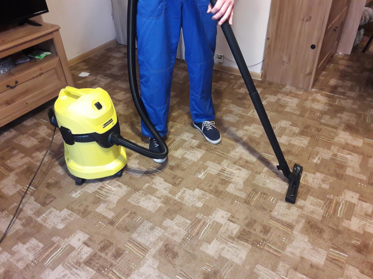 Osoba w niebieskim kombinezonie odkurza dywan żółtym odkurzaczem Karcher w pokoju z drewnianymi meblami.
