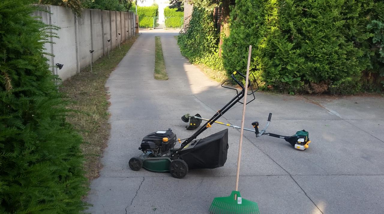 Kosiarka spalinowa, podkaszarka, trymer i grabie na betonowej nawierzchni podjazdu z pasem nieskoszonej trawy pośrodku, widok w kierunku bramy wjazdowej.