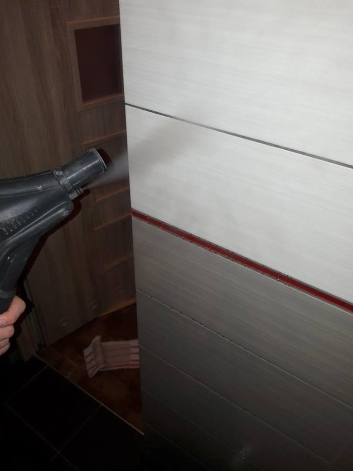 Czyszczenie parowe płytek ściennych w łazience, z użyciem parownicy ręcznej. Widoczna drewniana szafka i ściereczka na podłodze.