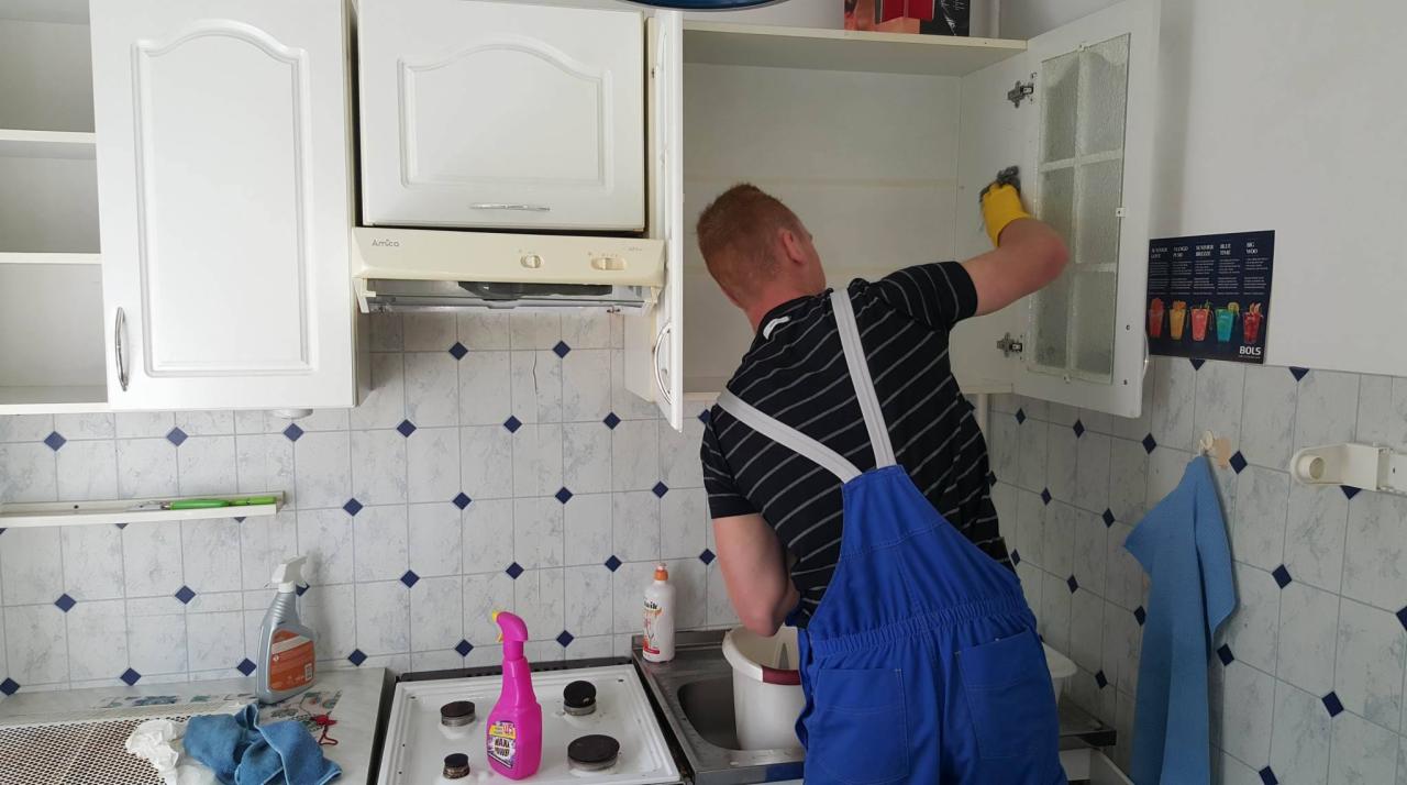 Mężczyzna w niebieskim kombinezonie i żółtych rękawicach czyści wnętrze szafki kuchennej z przeszklonymi drzwiczkami, widoczne środki czystości na blacie i kuchence.