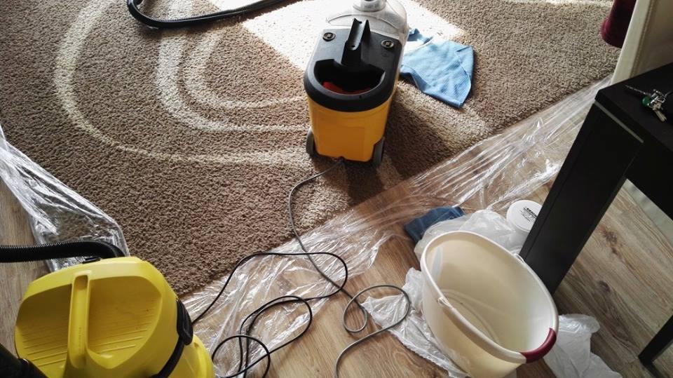 Żółty odkurzacz piorący na tle dywanu z okrągłym wzorem, zabezpieczonego folią ochronną, obok białe wiadro i niebieska ściereczka.