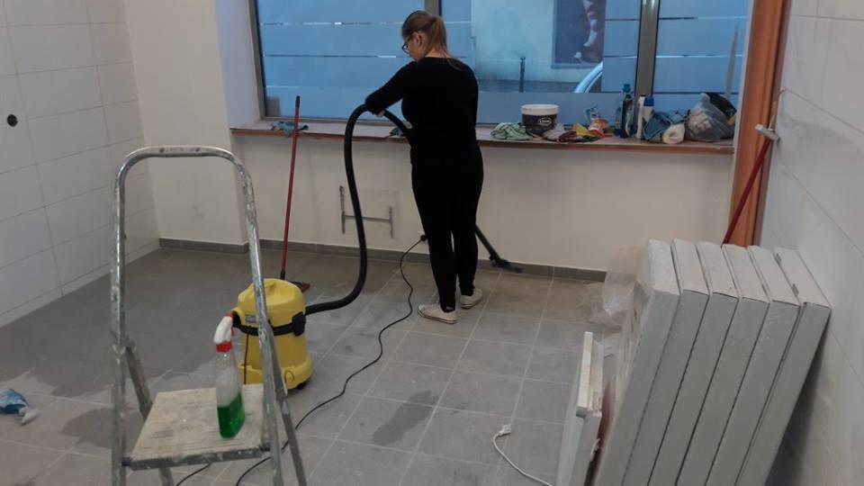 Kobieta w czarnym stroju odkurza szarą podłogę w pomieszczeniu z białymi płytkami, widoczny żółty odkurzacz, drabina, butelka ze środkiem czyszczącym i okno z akcesoriami na parapecie.