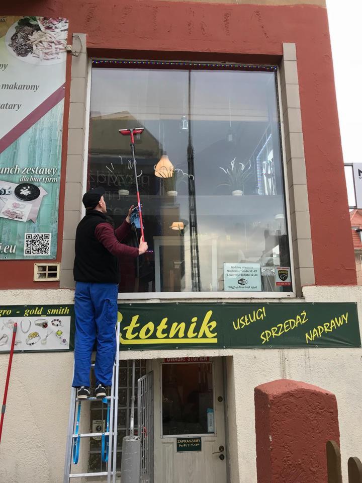Mężczyzna w czapce i niebieskich spodniach, stojący na aluminiowej drabinie, myje okno lokalu usługowego z zielonym szyldem 'Złotnik Usługi Sprzedaż Naprawy' za pomocą ściągaczki i szmatki.