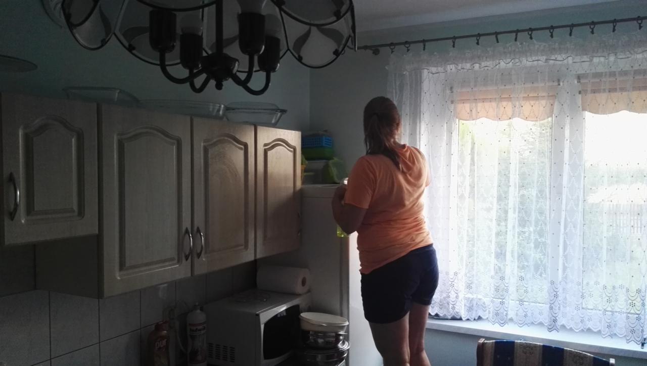 Kobieta stojąca na krześle, myjąca okno w kuchni z kremowymi szafkami i ozdobnymi zasłonami.