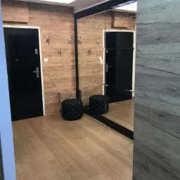 Wykończony przedpokój z panelami ściennymi imitującymi drewno, czarnymi drzwiami i lustrem z czarną ramą, uzupełniony o czarne pufy i wieszaki na ubrania.