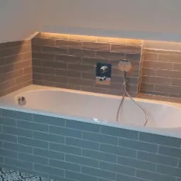 Wykończona łazienka na poddaszu z białą wanną obudowaną szarymi, podłużnymi płytkami, szarą glazurą na ścianach, podświetleniem LED w niszy nad wanną oraz dekoracyjną podłogą.