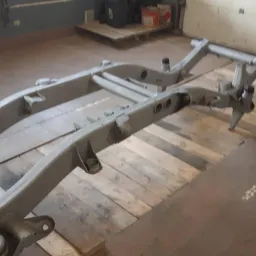 Pomarowana na szaro rama samochodu, leżąca na drewnianych paletach w warsztacie, widoczna z perspektywy lekko z góry.