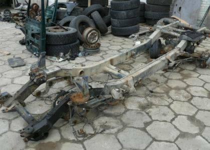 Zardzewiała rama samochodu z widocznymi elementami zawieszenia i silnika, otoczona stertą opon i części samochodowych na betonowej posadzce.